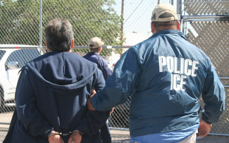 Este artículo habla sobre las prácticas de vigilancia de ICE reveladas en el informe American Dragnet. La imagen muestra a un oficial de ICE acompañando a un detenido.